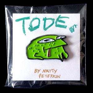 May include: Green enamel pin featuring a cartoon monster with a toothy grin and large eyes. The pin is packaged in a clear plastic bag with a white card that reads "TODE" and "BY NATTY PETERKIN".