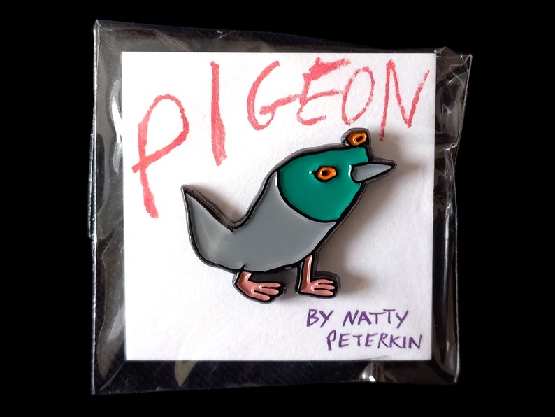 May include: A gray and green enamel pin of a cartoon pigeon with orange eyes and a black outline. The pin is on a white card with the word "PIGEON" written in red and the text "BY NATTY PETERKIN" in purple.