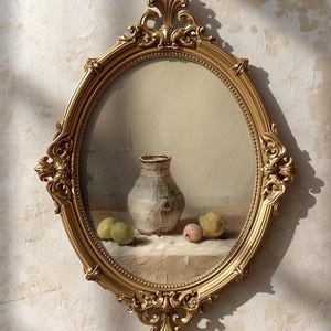Op de afbeelding: Ovale, sierlijke goudkleurige spiegel. Het kunstwerk van de spiegel toont een stilleven met een vaas en verschillende vruchten in groen- en roze tinten. De lijst heeft een gedetailleerd, vintage ontwerp.