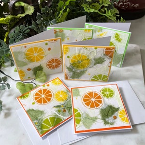 May include: Set of four blank greeting cards with a citrus fruit design. The cards feature a white background with a textured paper finish and a repeating pattern of orange and lime slices. The cards have orange and green borders.