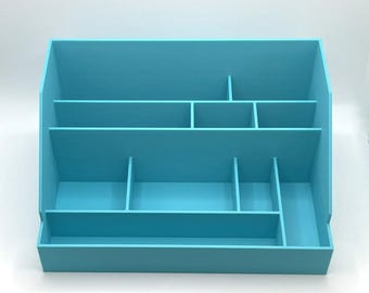 May include: A light blue desk organizer with multiple compartments and shelves. The organizer has a rectangular base and a sloped back, with various sized sections for storage. The color is a solid, bright blue.