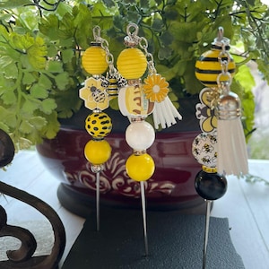 May include: Three bee-themed decorative pins with yellow, black, and white beads. The pins are displayed on a black foam block.