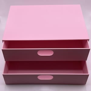 Peut inclure: Une boîte de rangement à deux tiroirs dans les tons de rose. Le dessus et les tiroirs sont d'un rose plus clair, tandis que l'intérieur des tiroirs est d'un rose plus foncé. Chaque tiroir a une poignée ovale arrondie.