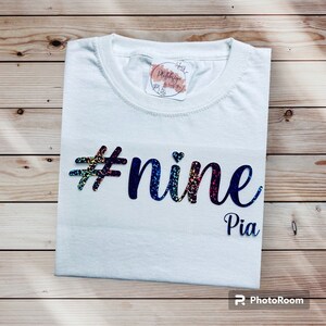 Könnte beinhalten: Ein weißes T-Shirt mit dem Text "#nine Pia" in einer bunten, glitzernden Schrift. Das Shirt ist sauber auf einer Holzoberfläche gefaltet. Ein kleiner Aufkleber ist in der Nähe des Kragens zu sehen.