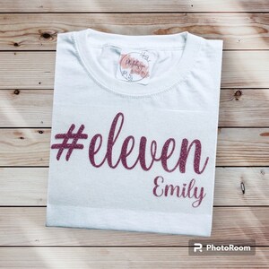 Könnte beinhalten: Weißes T-Shirt mit Rundhalsausschnitt. Auf dem Shirt steht in glitzerndem Burgunderrot "#eleven" und darunter der Name "Emily". Das Shirt liegt auf einer Holzoberfläche.