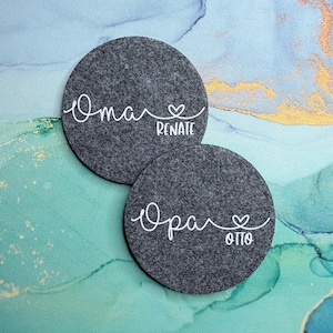 Könnte beinhalten: Zwei dunkelgraue Filzuntersetzer mit weißem Schreibschrifttext. Einer der Untersetzer trägt die Aufschrift "Oma Renate" mit einem Herzsymbol. Der andere trägt die Aufschrift "Opa Otto" mit einem Herzsymbol. Die Untersetzer sind kreisförmig.