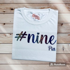 Könnte beinhalten: Ein weißes T-Shirt mit dem Text "#nine Pia" in einer bunten, glitzernden Schrift. Das Shirt ist sauber auf einer Holzoberfläche gefaltet. Ein kleiner Aufkleber ist in der Nähe des Kragens zu sehen.