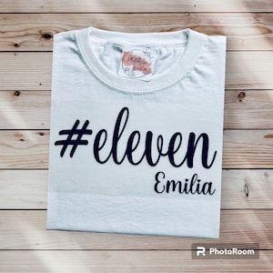 Könnte beinhalten: Weißes T-Shirt mit dem Text "#eleven Emilia" in dunkelblauer Schrift. Das T-Shirt ist auf einer hellbraunen Holzoberfläche gefaltet. Auf dem Kragen befindet sich ein kleiner Aufkleber mit Text und floralen Designs.