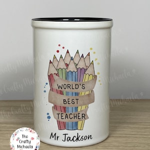 May include: A white ceramic pencil holder with a black rim featuring a watercolour illustration of coloured pencils wrapped in a tan ribbon. The ribbon displays the text "WORLD'S BEST TEACHER" in black, with "Mr Jackson" printed below.