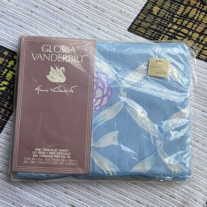 May include: A blue twin flat sheet with a white floral pattern and a white ribbon design. The sheet is packaged in plastic and has a label that reads "Gloria Vanderbilt" and "One Twin Flat Sheet, No Iron, Fine Percale, 200 Threads Per Sq. In., For 39 x 75in Mattress (99 x 190cm), 66 x 96in Finished Size (167 x 243cm)".