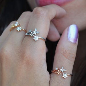 Puede incluir: Tres anillos de plata con un delicado diseño floral con pequeñas piedras de circonita cúbica transparentes. Los anillos son ajustables y se pueden llevar en diferentes dedos.