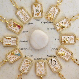 May include: A set of 12 gold zodiac necklaces with white and gold pendants. Each pendant features a different zodiac sign with a white background and gold accents. The signs are: Aries, Taurus, Gemini, Cancer, Leo, Virgo, Libra, Scorpio, Sagittarius, Capricorn, Aquarius, and Pisces.
