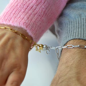 May include: Gold and silver chain bracelets with heart-shaped charms and initial pendants. One bracelet is gold, the other is silver. The charms include the letters "M" and "C". The bracelets are connected by a magnetic clasp.