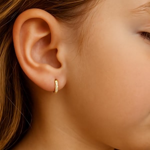 Puede incluir: Primer plano de un pendiente de aro dorado con pequeñas piedras transparentes en la parte delantera. El pendiente se lleva en una oreja, con un tono de piel claro y cálido. Una pieza de joyería clásica.