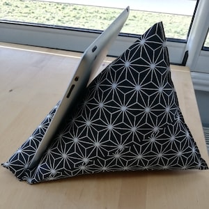 Peut inclure: Un support de tablette en forme de coussin avec un motif géométrique noir et blanc. Le coussin triangulaire soutient une tablette argentée. Le support est en tissu et repose sur une surface en bois brun clair.