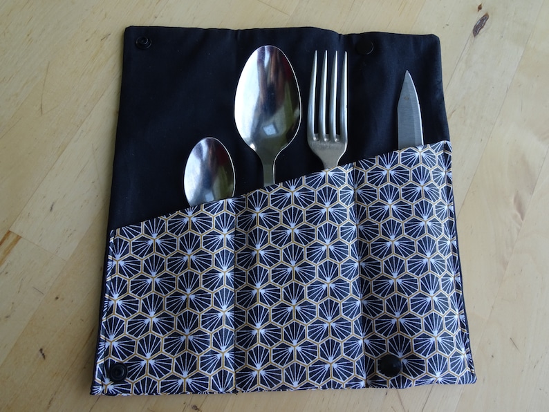 May include: A black fabric cutlery roll with a geometric pattern in gold and white. The roll has three slots for utensils, and a knife, fork, and two spoons are visible.