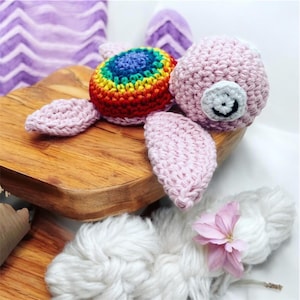 Peut inclure: Un jouet en crochet en forme de tortue rose et arc-en-ciel avec un visage souriant. La tortue est posée sur une surface en bois.