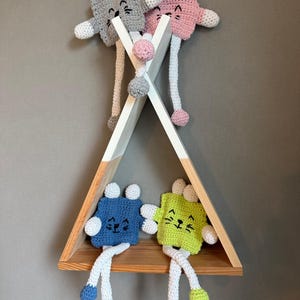 Könnte beinhalten: Ein handgefertigtes Holzregal in Form eines Tipi, mit vier gehäkelten Katzenpuppen. Die Puppen sind in Grau-, Rosa-, Blau- und Grüntönen gehalten, mit weißen und farbigen Akzenten. Das Regal ist weiß mit einem Regal aus Naturholz.
