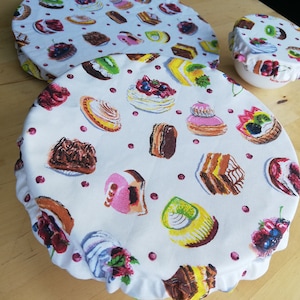 Op de afbeelding: Witte stoffen komhoezen met een kleurrijk patroon van diverse gebakjes, taarten en bessen. De hoezen zijn in verschillende maten, met een geschulpte rand op de grootste. Een kleine kom is bedekt met een bijpassend deksel.