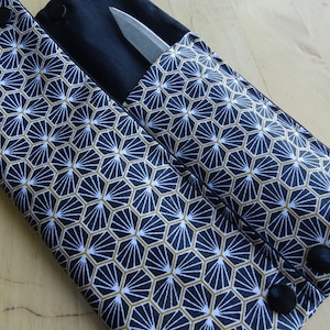 May include: A black fabric knife roll with a geometric pattern in gold and white. The roll has two pockets and two black buttons.