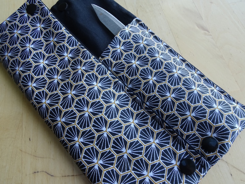 May include: A black fabric knife roll with a geometric pattern in gold and white. The roll has two pockets and two black buttons.