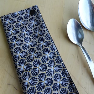 May include: A black fabric cutlery holder with a geometric pattern in gold and white. The holder is closed with a black button. Two silver spoons are lying on a wooden surface.