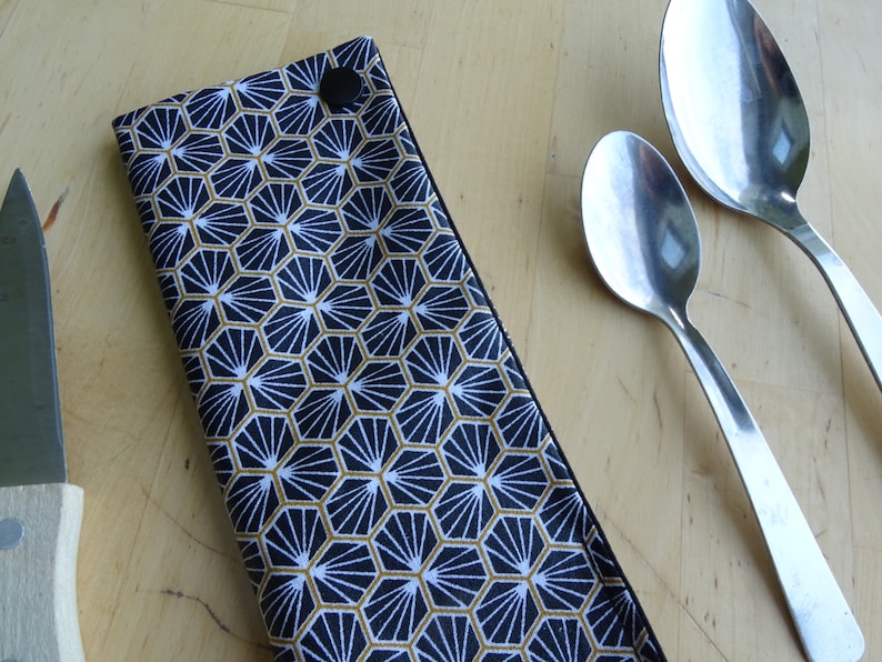 May include: A black fabric cutlery holder with a geometric pattern in gold and white. The holder is closed with a black button. Two silver spoons are lying on a wooden surface.