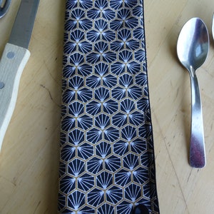 May include: A black and gold fabric cutlery holder with a geometric pattern. The holder has two black buttons on the sides.