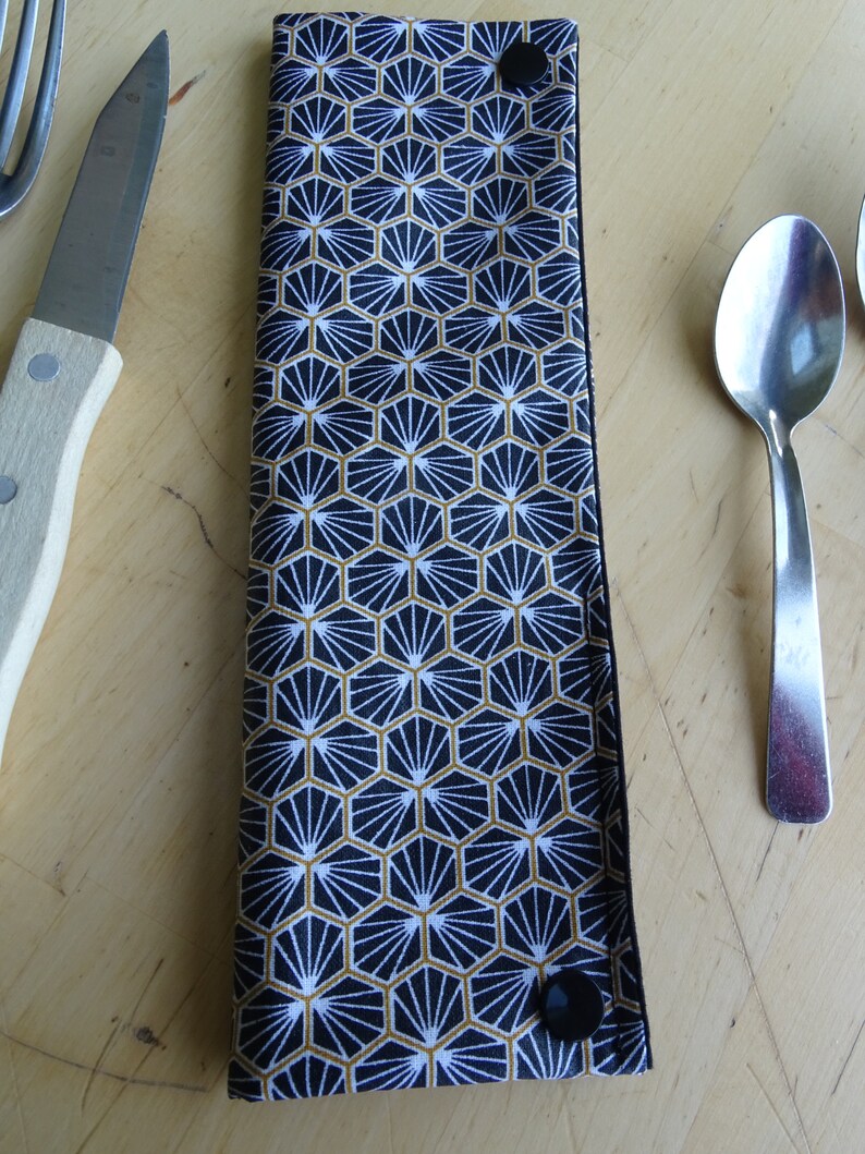 May include: A black and gold fabric cutlery holder with a geometric pattern. The holder has two black buttons on the sides.