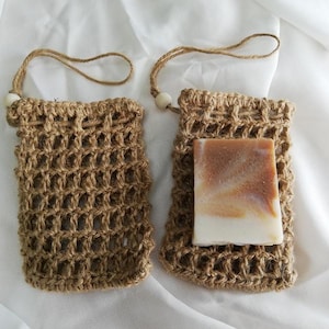 May include: Two woven jute soap savers with a natural bar of soap. The soap saver has a loop for hanging and a wooden bead detail. The soap is a light brown and white color.
