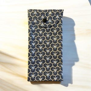 Pode incluir: Uma bolsa retangular de tecido com fundo preto e um padrão geométrico hexagonal repetitivo em branco e dourado. A bolsa possui um fecho de aba pontiaguda fixado com um único botão de pressão preto centralizado na parte frontal.