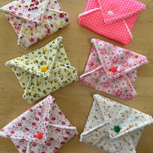 May include: Six reusable cloth sanitary pads in floral and polka dot designs. The pads are square, folded, and secured with a button. Colours include pink, yellow, and white with floral and polka dot patterns. Each pad has a white trim.