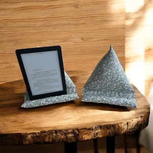 Op de afbeelding: Een e-reader rust op een kussenstandaard met bloemenprint, naast een bijpassend driehoekig kussen. De e-reader toont tekst op het scherm. De kussens zijn lichtgroen met een wit bloemmotief, op een houten tafel.