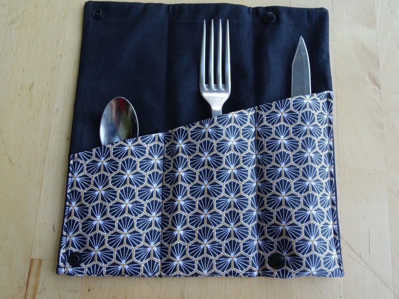 May include: A black fabric cutlery roll with a geometric pattern in white and gold. The roll has three slots for a fork, spoon, and knife.