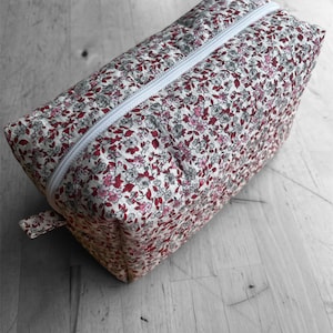 Puede incluir: Un neceser rectangular con estampado floral y cremallera blanca. La bolsa presenta un patrón de flores rojas, rosas y grises sobre un fondo crema. La cremallera se extiende por la parte superior.