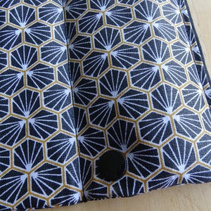 May include: A black and white fabric with a geometric pattern of gold hexagons with a white fan design inside each hexagon. The fabric has a black button in the lower right corner.