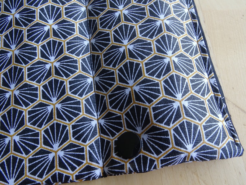 May include: A black and white fabric with a geometric pattern of gold hexagons with a white fan design inside each hexagon. The fabric has a black button in the lower right corner.