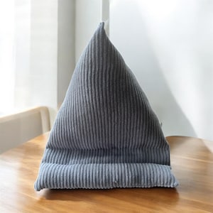 May include: A gray corduroy triangular pillow, designed to prop up books or tablets. The pillow has a ridged texture and is placed on a wooden surface. The pillow's shape is reminiscent of a mountain.