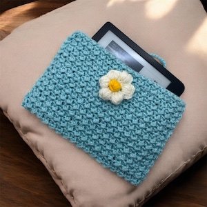 Può includere: Una pochette in crochet blu chiaro con un fiorellino di margherita bianco come decorazione. La pochette è progettata per contenere un e-reader.