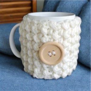 Op de afbeelding: Een witte keramische mok met een crèmekleurige, handgebreide hoes. De hoes is vastgezet met een grote, lichtbruine houten knoop. De mok staat op een blauw oppervlak, wat wijst op een gezellig, handgemaakt thema.