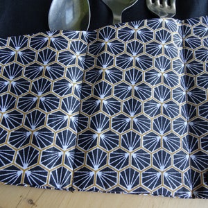 May include: A black fabric cutlery roll with a geometric pattern in white and gold. The roll has two black buttons and is designed to hold silverware.