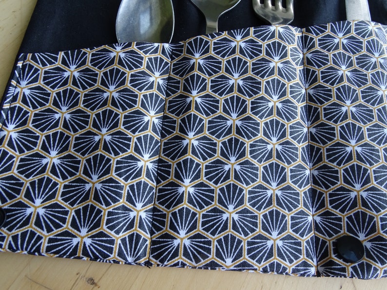May include: A black fabric cutlery roll with a geometric pattern in white and gold. The roll has two black buttons and is designed to hold silverware.