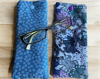 May include: Two fabric eyeglass cases with different patterns. One case is blue with a small white flower pattern. The other case is blue, grey, and purple with a floral pattern. Both cases have a button closure.