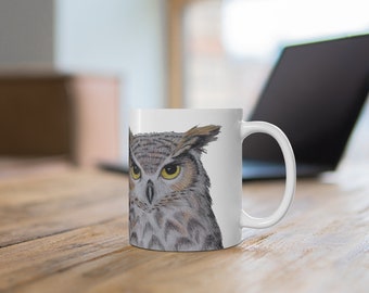 May include: White ceramic mug featuring a close-up image of an owl's face. The owl has brown and grey feathers and yellow eyes.