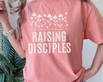 May include: A coral-colored t-shirt with the words "RAISING DISCIPLES" in white block letters. Above the text is a white floral design. The shirt is a comfortable, casual style.