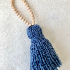 May include: A blue tassel with a wooden bead garland attached. The tassel is made of soft yarn and has a long, flowing fringe.