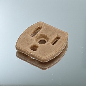 Puede incluir: Una pieza de pl&aacute;stico beige impresa en 3D con un agujero circular y tres recortes rectangulares.
