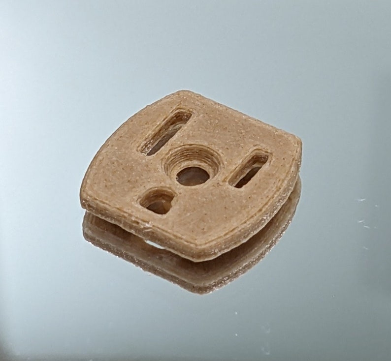 Puede incluir: Una pieza de pl&aacute;stico beige impresa en 3D con un agujero circular y tres recortes rectangulares.