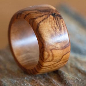 May include: A wide band ring crafted from polished olive wood, showcasing a natural light and dark brown grain pattern with a smooth, rounded exterior surface and a hollow center, highlighting the organic texture of the wood material.