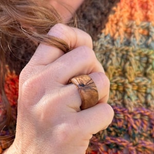 May include: A close-up of a wooden ring with a natural wood grain pattern. The ring is worn on a person's finger.